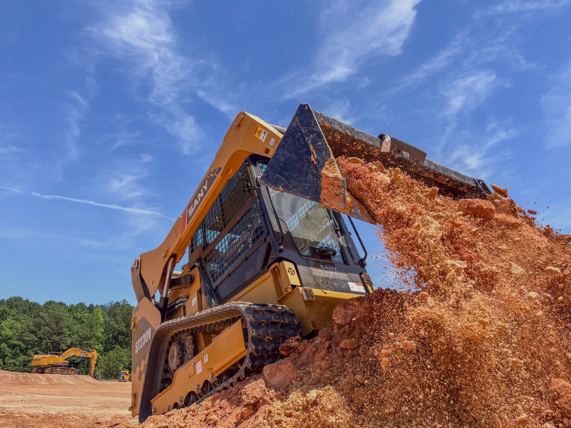 A yellow compact track loader dumping a bucket of red dirt on a sunny day.