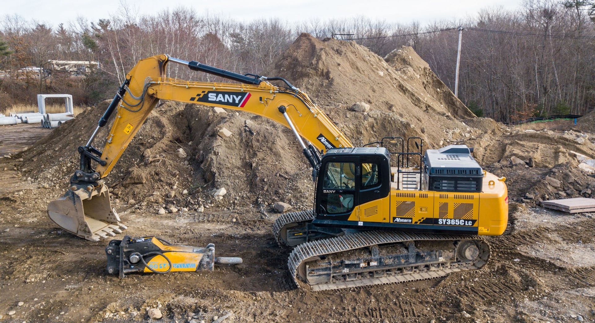 A yellow SANY excavator sits on a dirt construction site with a hydraulic hammer attachment on the ground in front of it.