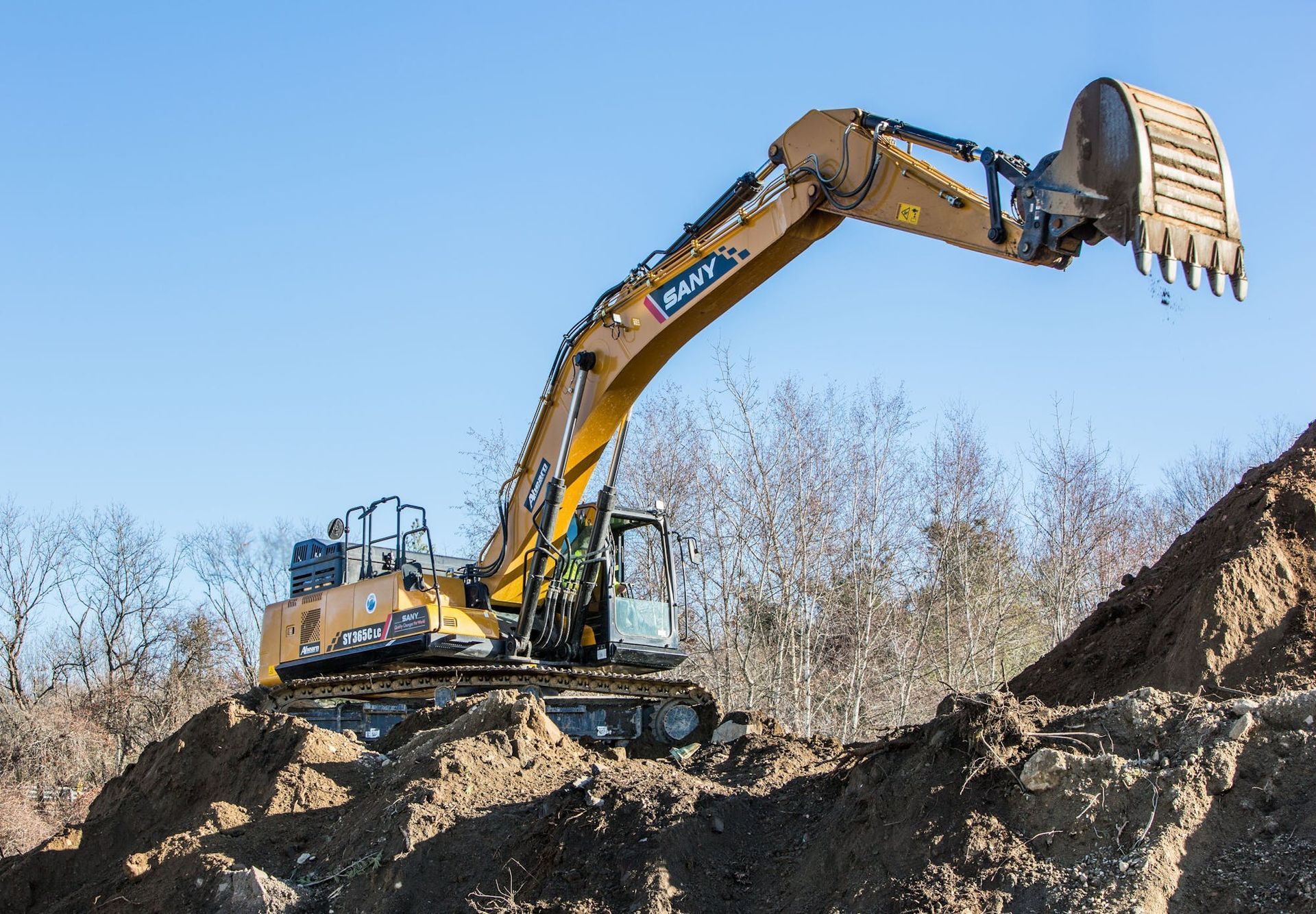 A yellow Sany excavator with its bucket raised, positioned on a dirt mound against a clear blue sky.