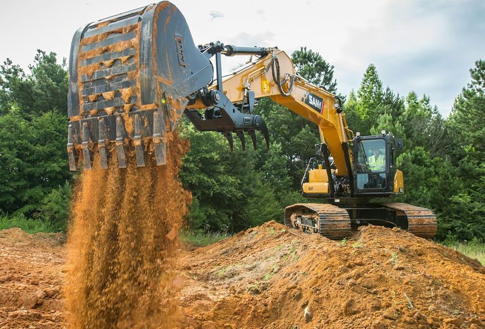 Yellow excavator dumping a load of brown soil onto a pile. Trees and sky in background.