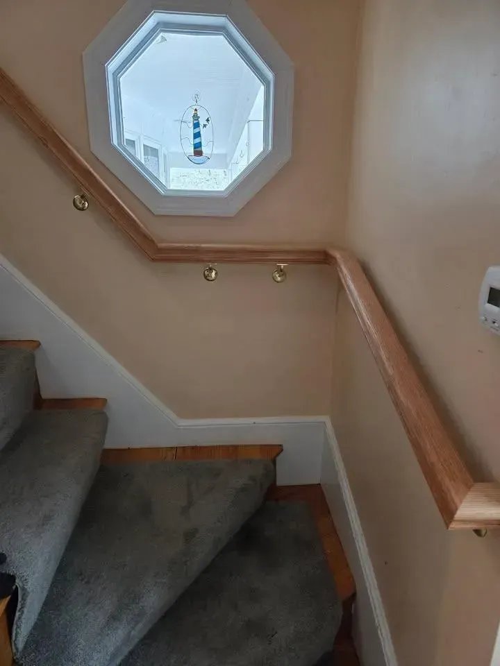 Staircase landing with beige walls, gray carpet, wooden handrails, and a small octagonal window.