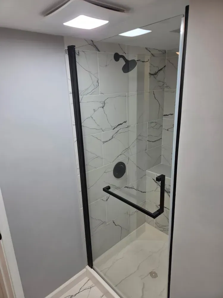 Modern shower stall with marble-look tile, black fixtures, and a glass door under bright ceiling lights