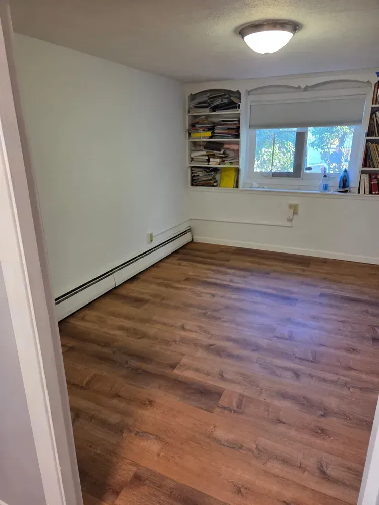 Empty bedroom with wood flooring, white walls, built-in shelves, and a window with blinds