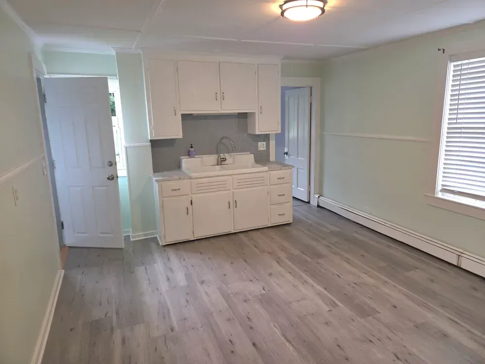 Empty studio apartment with white kitchen cabinets, gray flooring, and a window with blinds