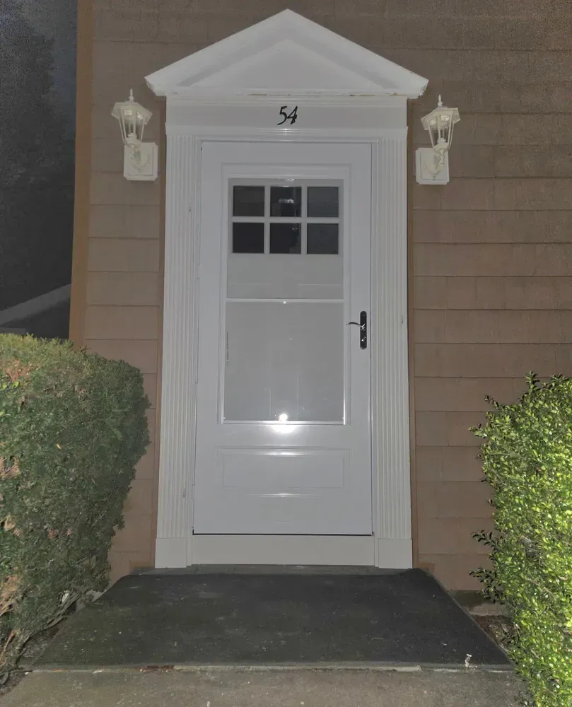 Front door with white trim, small window, and house number 54 beside bushes at night