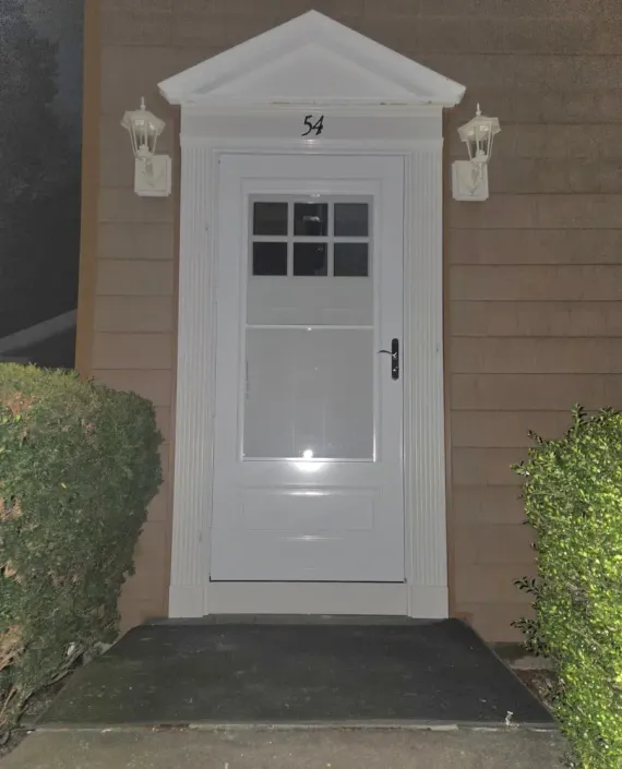 Front door with white trim, small window, and house number 54 beside bushes at night