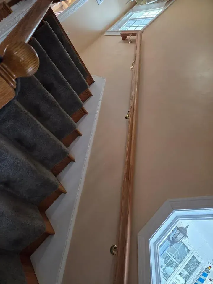Staircase with dark carpeted steps, beige wall, wooden handrail, and window light.