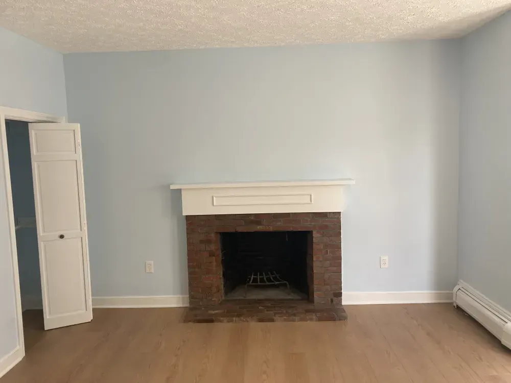 Empty living room with light blue walls, brick fireplace, white mantel, and beige carpet