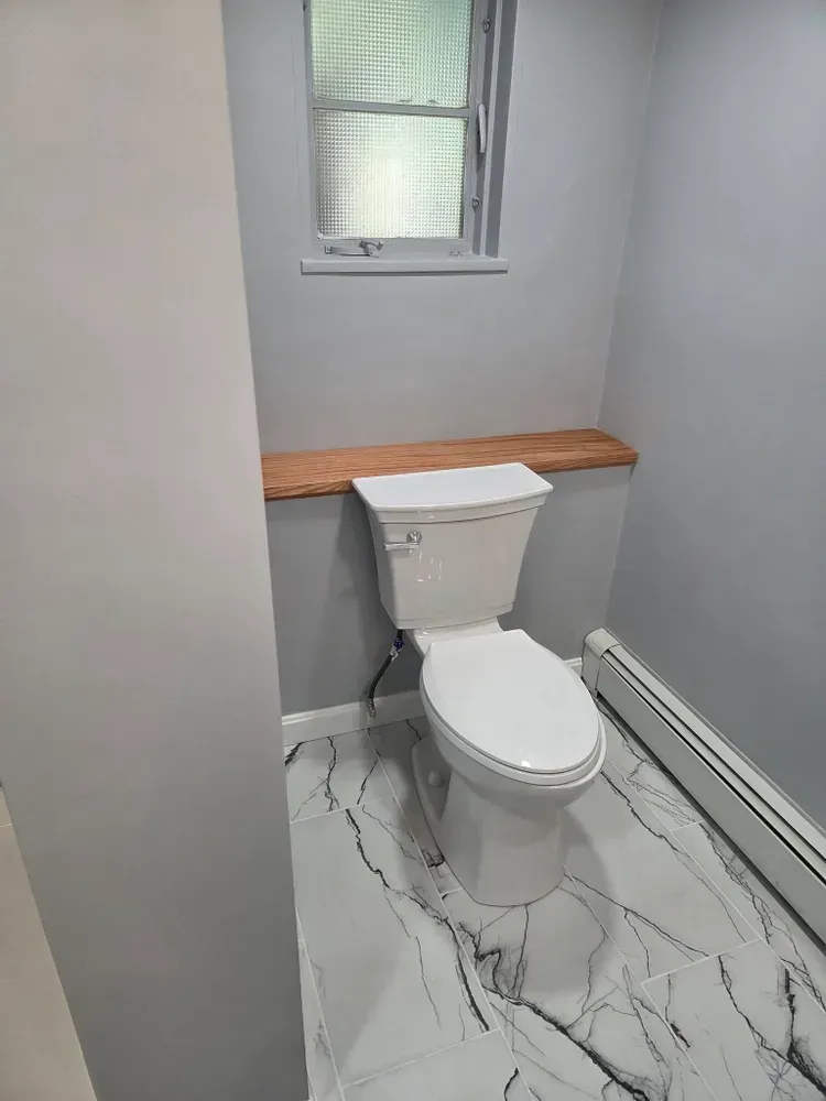 Small bathroom with a toilet, wooden shelf, frosted window, and gray walls with marble-patterned floor.