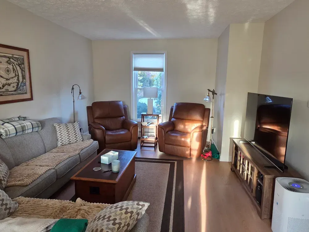 Bright living room with sofa, two leather chairs, TV, coffee table, and sunlight by the patio door
