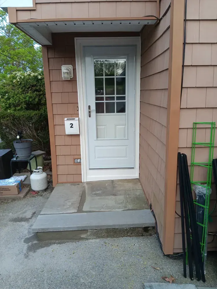 Front entrance with white door, concrete steps, tan siding, and green folded chairs by the wall.