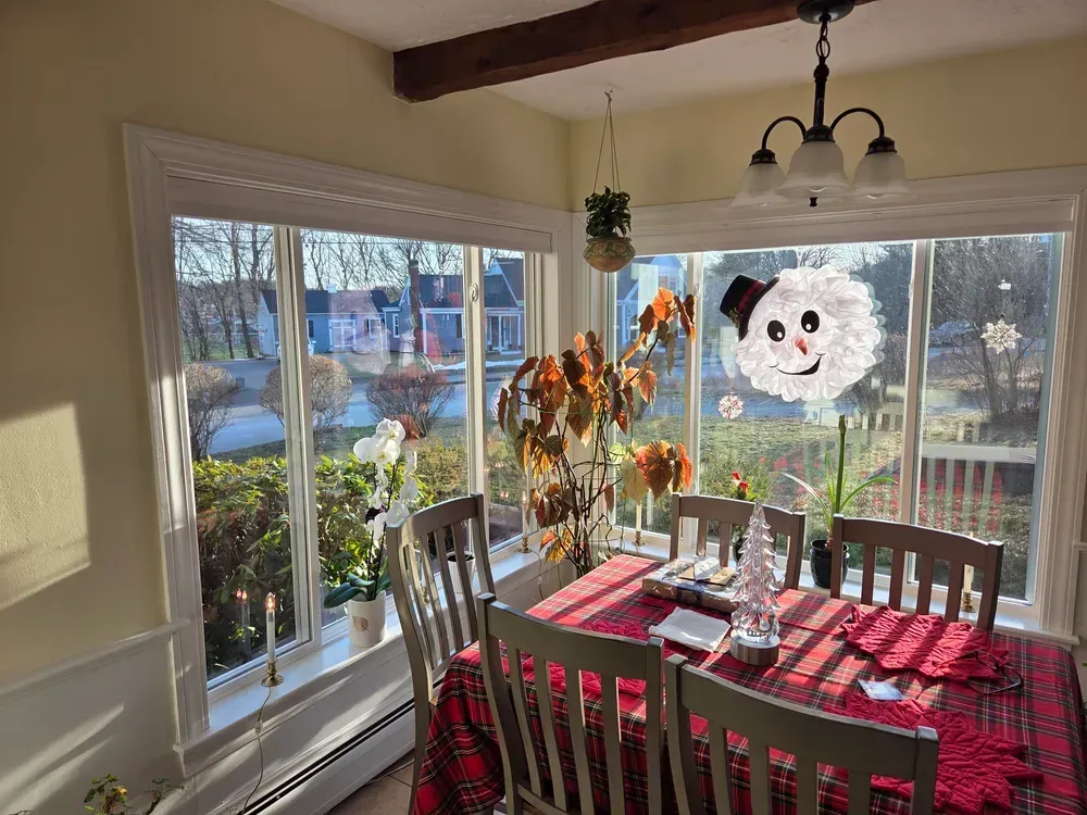 Sunlit dining room with red table runner, wooden table, chairs, and autumn wreath on window overlooking water