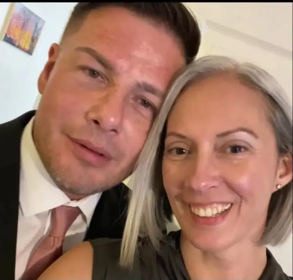 Two smiling people posing closely for a selfie indoors, one in a suit and tie.