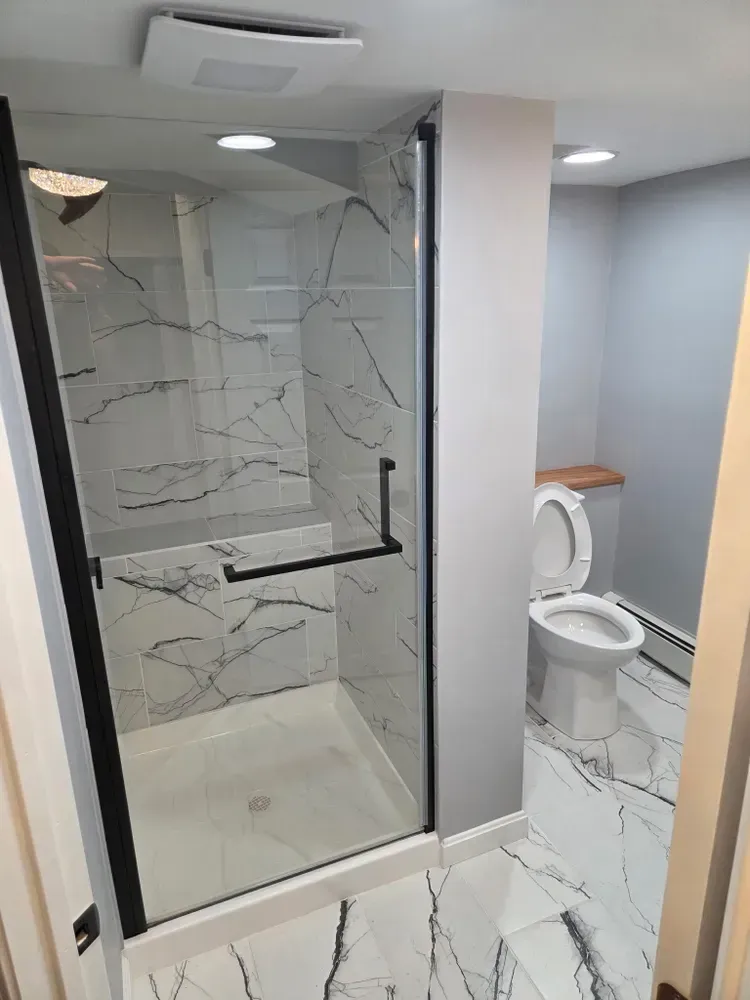Modern bathroom with glass shower, white marble tile, and a toilet beside a vanity.