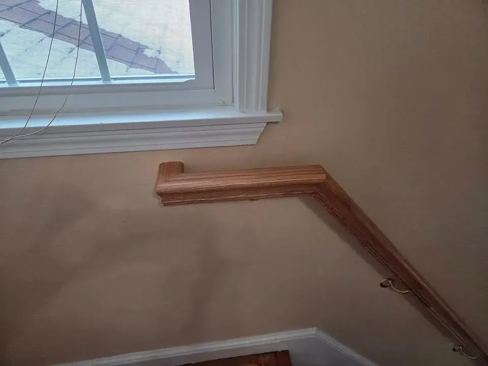 Wooden handrail mounted along a stairway wall beneath a window, with a damaged or unfinished section near the top.