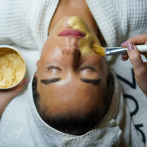 Person receiving a gold facial mask. Face is up, eyes closed. Hand applies mask with a brush. Jar of mask nearby.