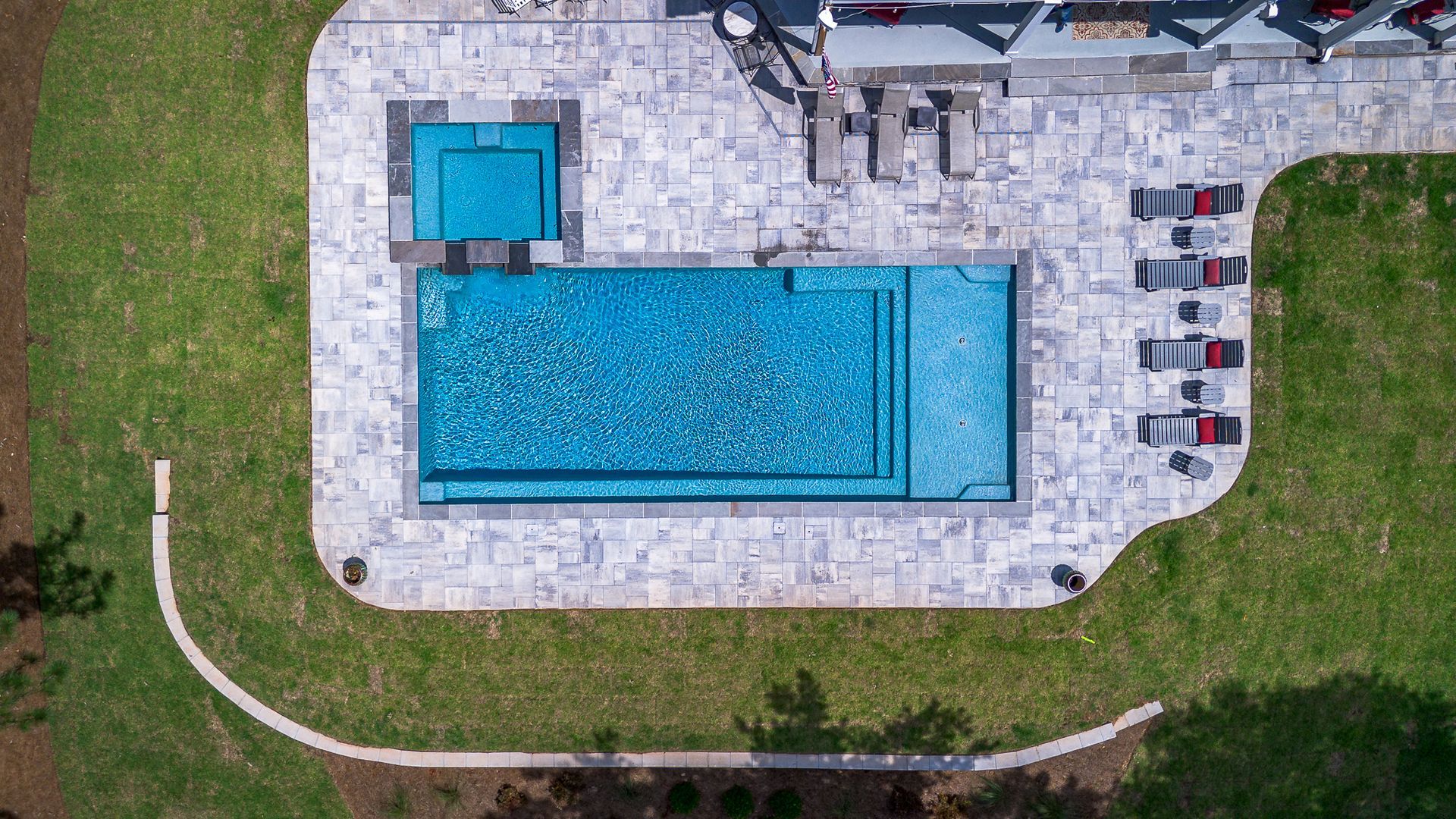 Aerial view of a pool with a hot tub, surrounded by stone patio and lawn.