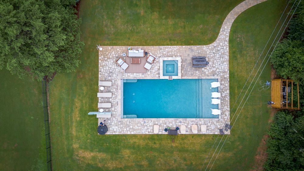 Aerial view of a pool surrounded by a stone patio with lounge chairs, a hot tub, and green grass.