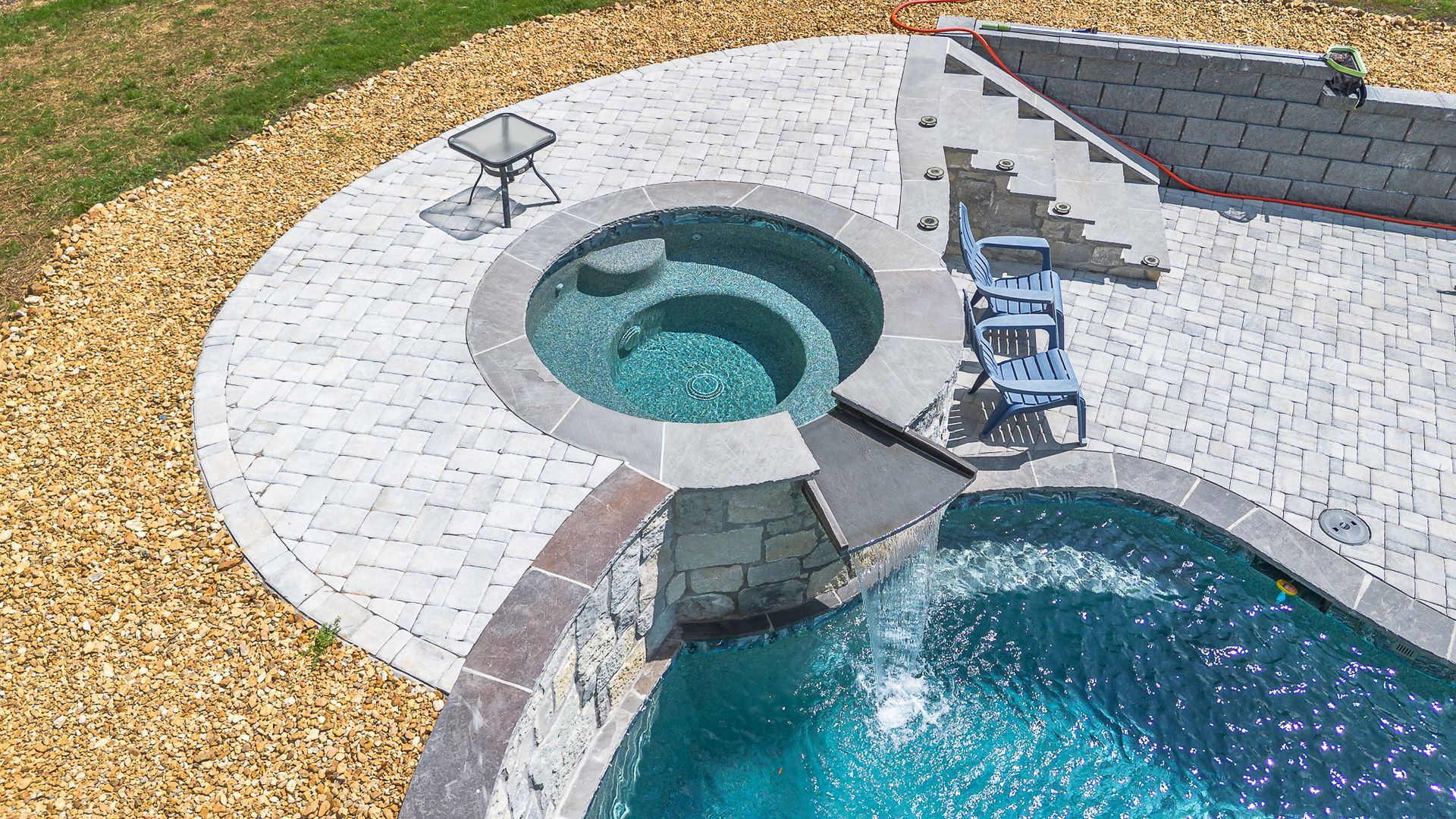 Overhead view of a pool and spa, with a stone patio, flowing water, and lounge chairs.