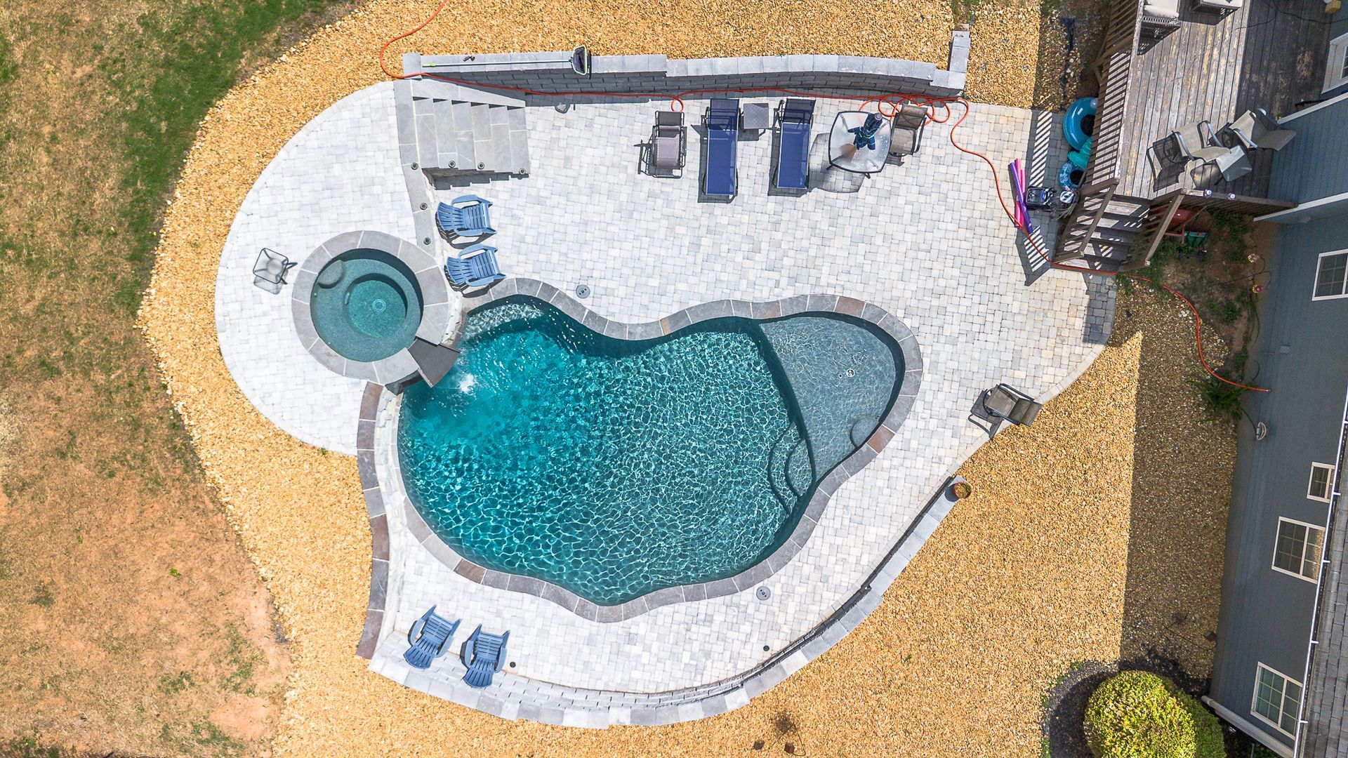 Overhead view of a pool with a spa, surrounded by light-colored stone patio and gravel landscaping.