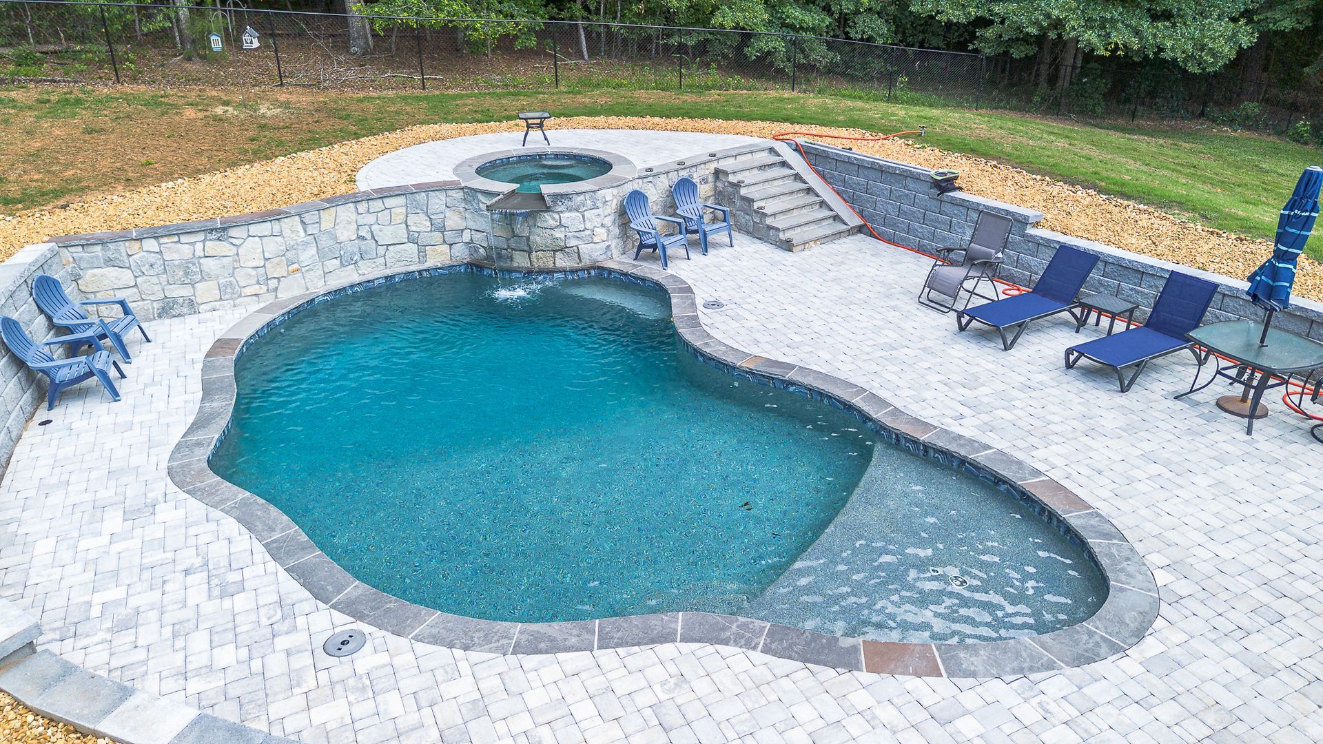 Pool with spa, waterfall, and patio area with chairs. Blue water, stone accents, outdoor setting.