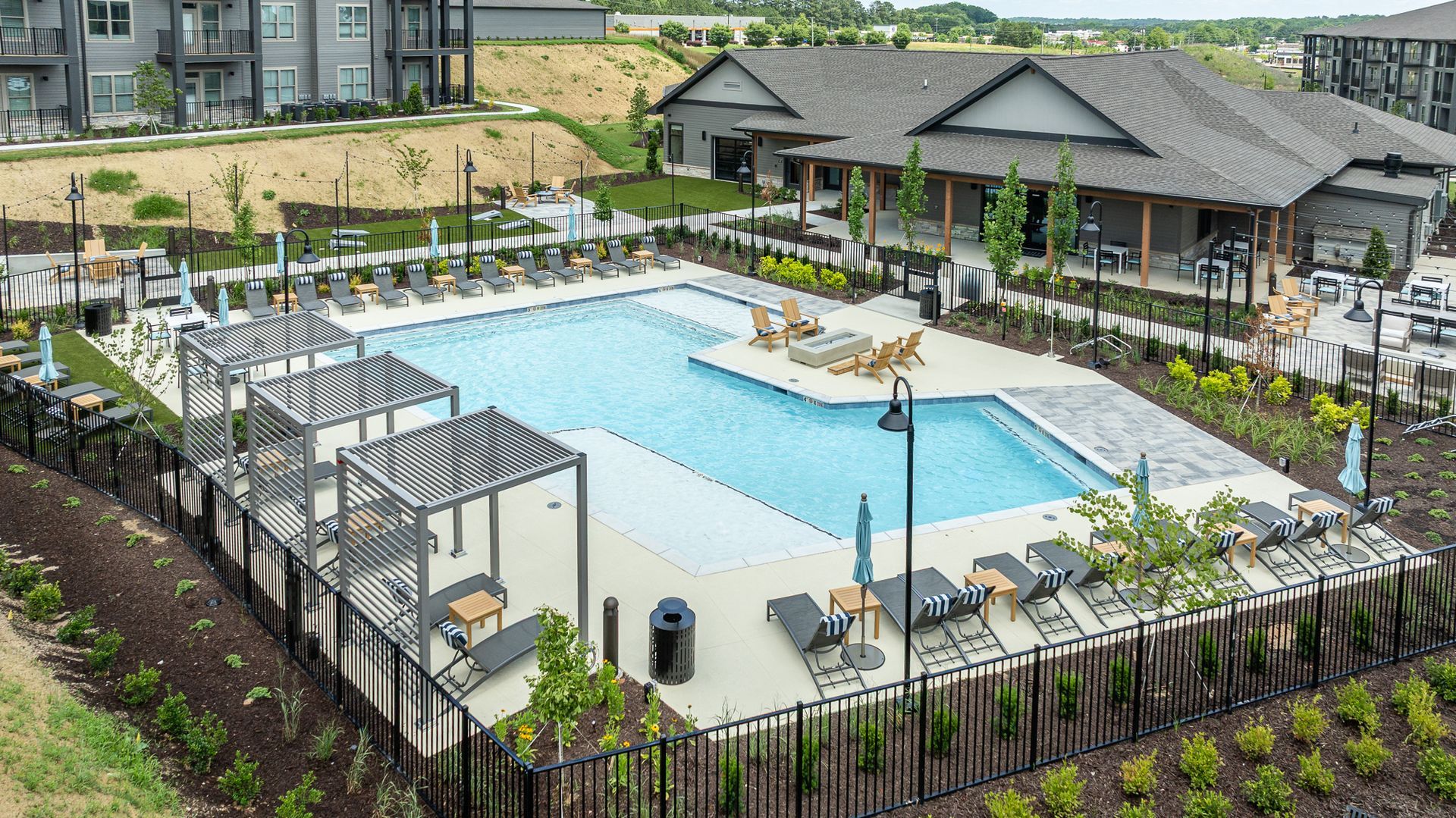 Aerial view of a modern apartment pool with lounge chairs, fenced deck, and clubhouse building