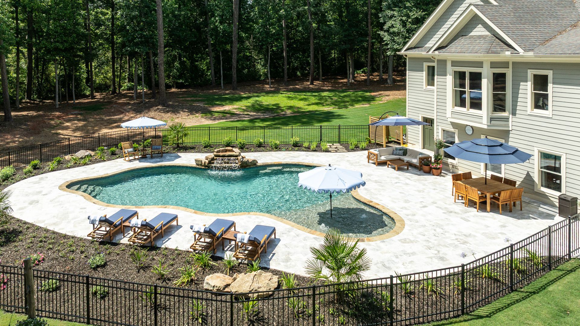 Backyard pool with patio seating beside a house, surrounded by trees and a black fence