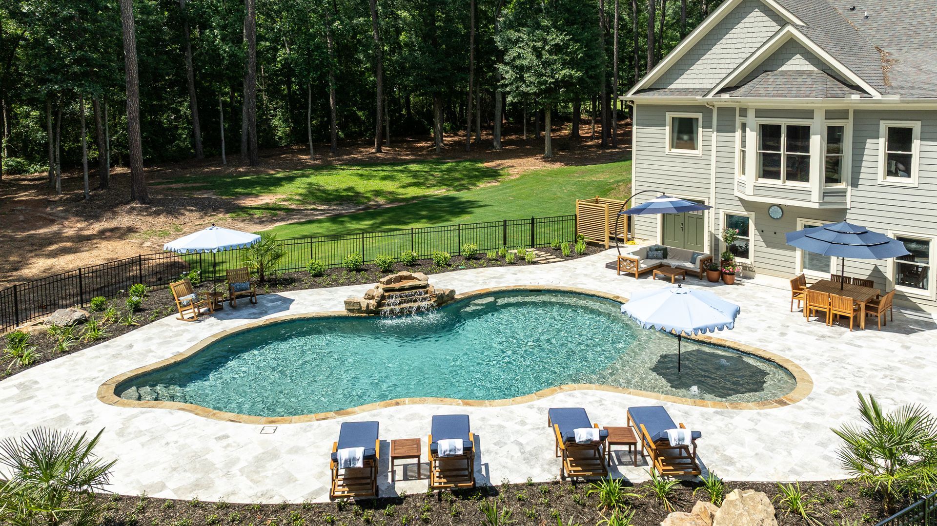 Backyard pool with patio chairs and tables beside a house, surrounded by trees and a landscaped yard