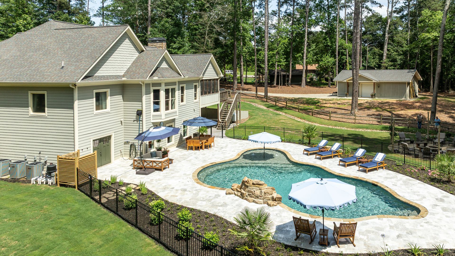 Backyard patio with a pool, lounge chairs, umbrellas, and a house beside a wooded area