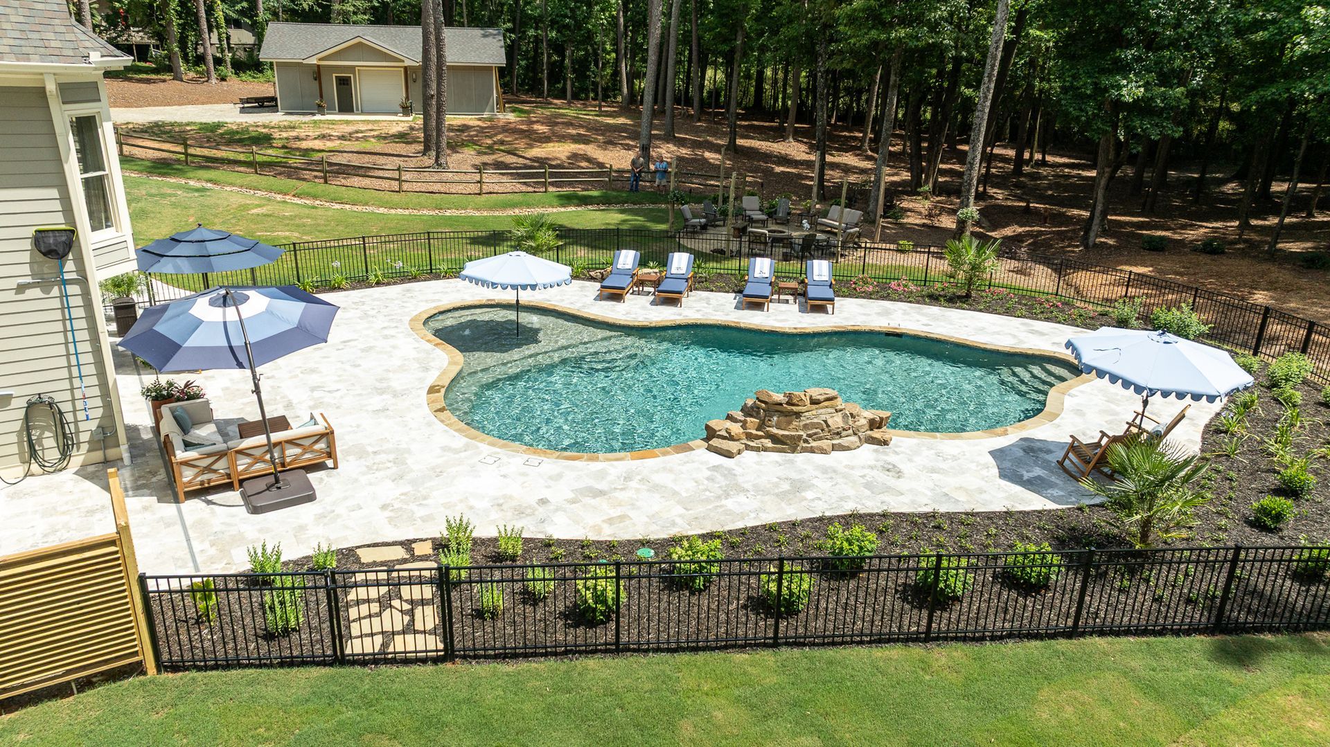 Backyard pool with stone patio, lounge chairs, umbrellas, and a small rock waterfall, bordered by trees.