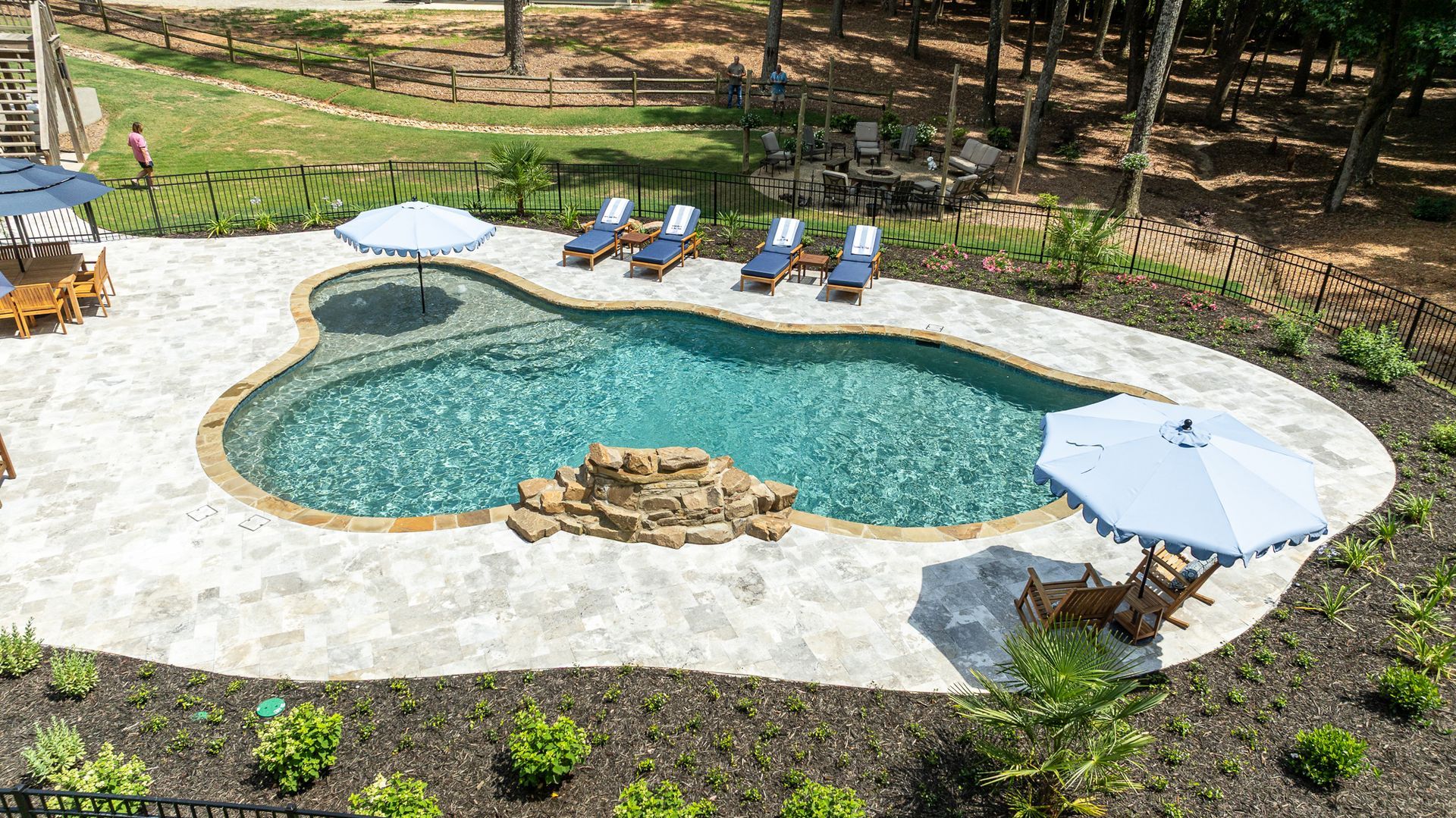 Backyard pool with curved stone deck, lounge chairs, and umbrellas surrounded by greenery