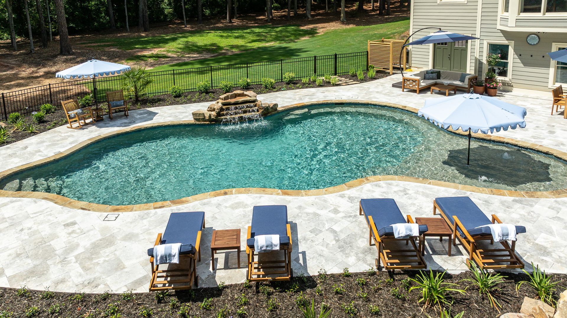Backyard pool with lounge chairs, umbrellas, and a small waterfall surrounded by trees