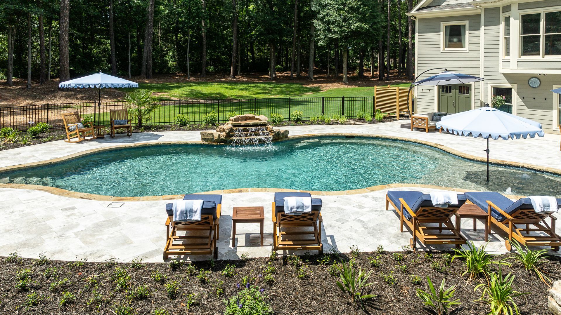 Backyard pool with stone waterfall, lounge chairs, and white patio umbrellas beside a house and trees