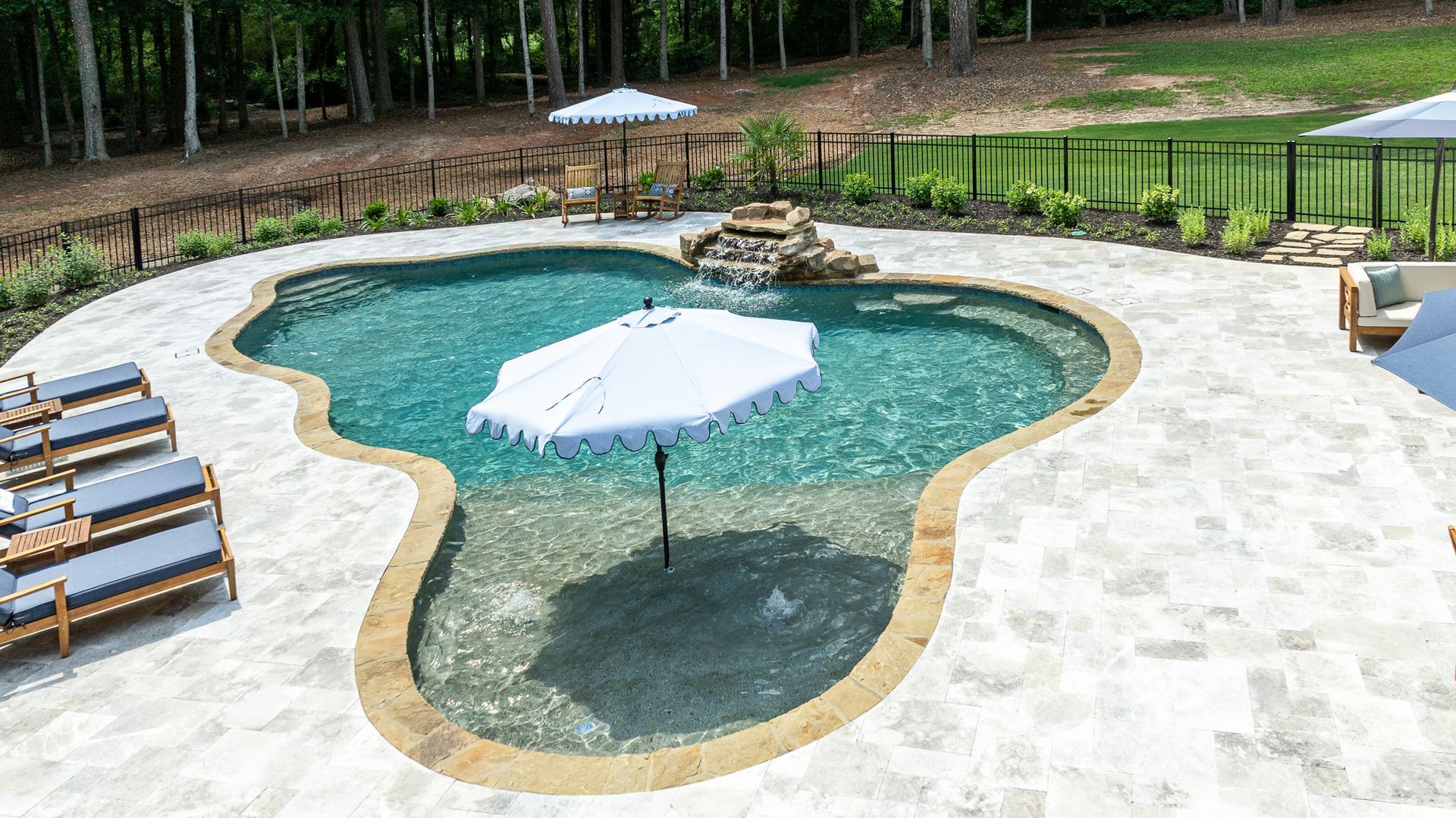 Outdoor swimming pool with turquoise water, stone deck, lounge chairs, and umbrellas beside a wooded area