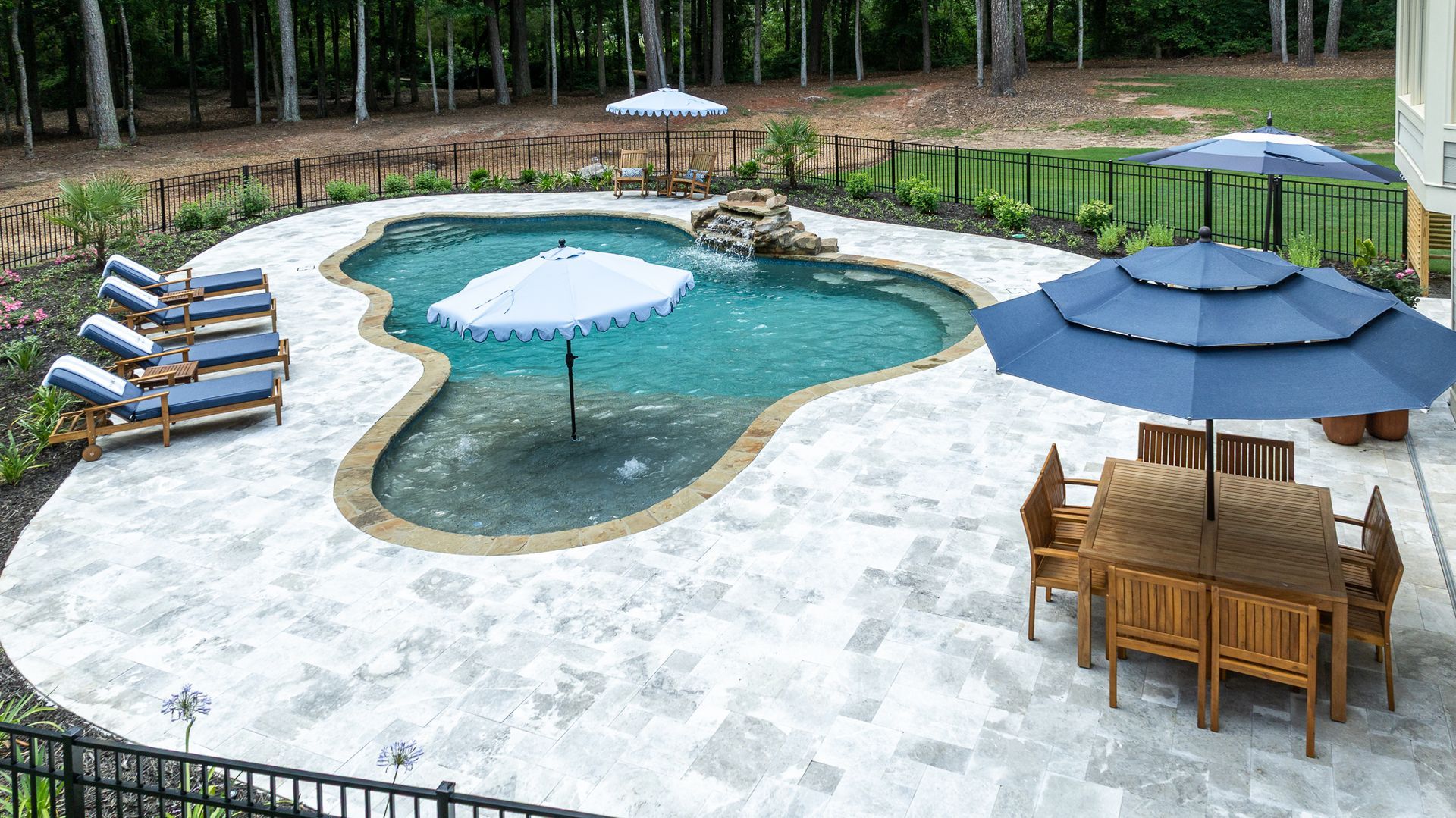 Backyard pool with stone deck, lounge chairs, and blue patio umbrellas amid trees