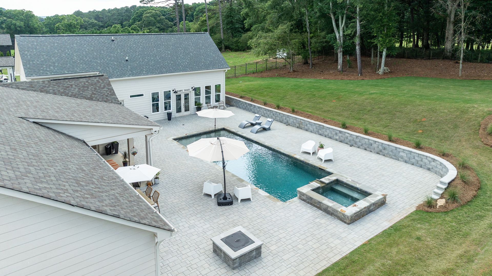 Backyard patio with rectangular pool, hot tub, umbrella seating, and a white house beside a grassy yard