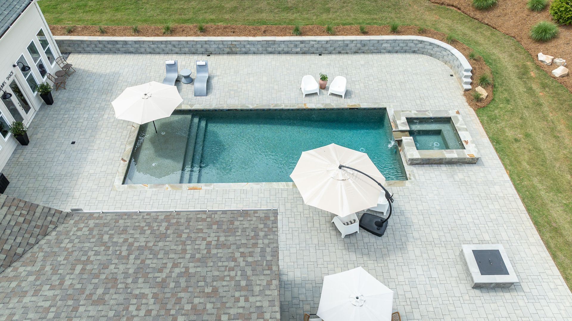 Overhead view of a backyard pool with patio umbrellas, lounge chairs, and a small hot tub.