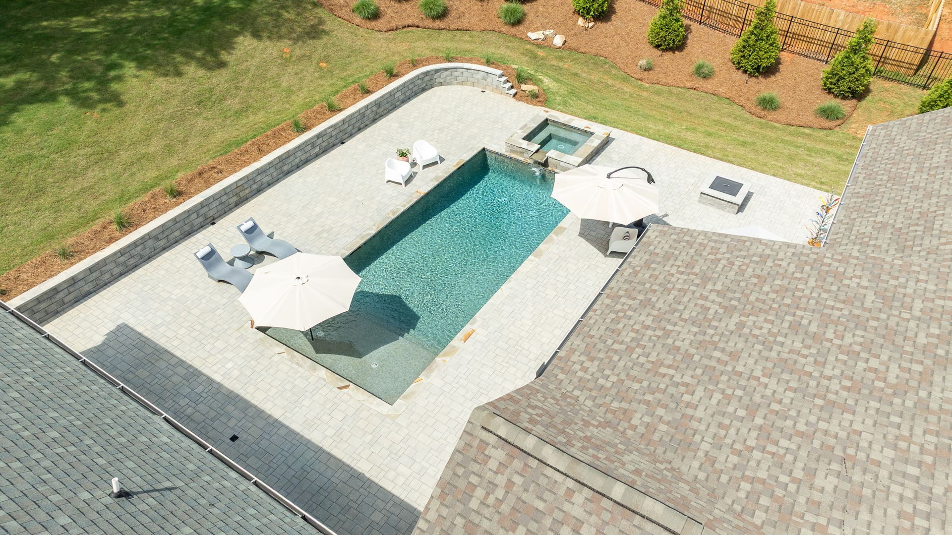 Aerial view of a backyard pool with white lounge chairs, umbrellas, and a paved patio surrounded by grass.