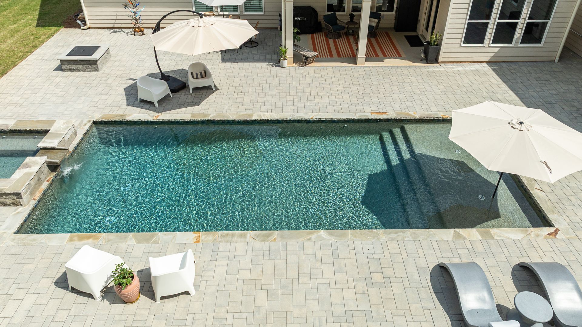 Aerial view of a backyard pool with white lounge chairs, umbrellas, and a patio beside a house