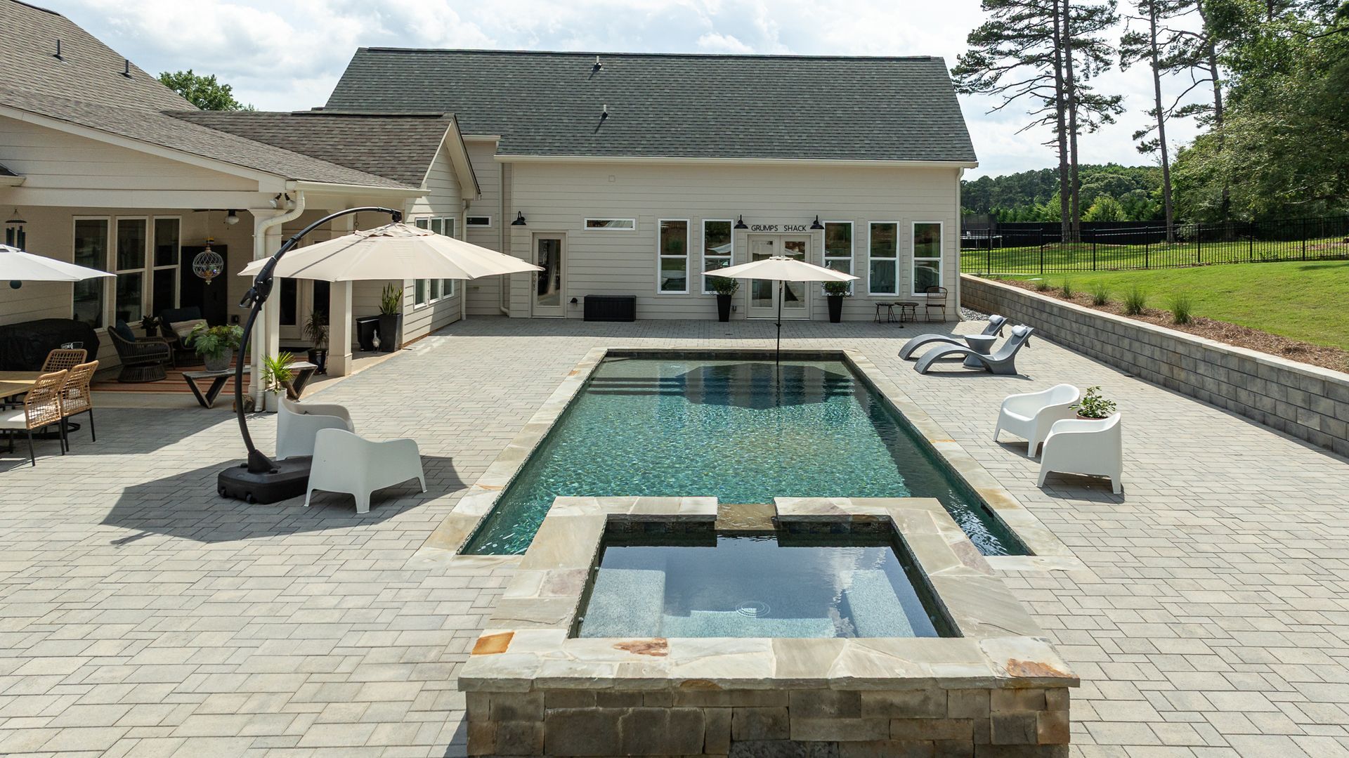 Backyard pool with hot tub, loungers, and patio beside a white house under clear skies