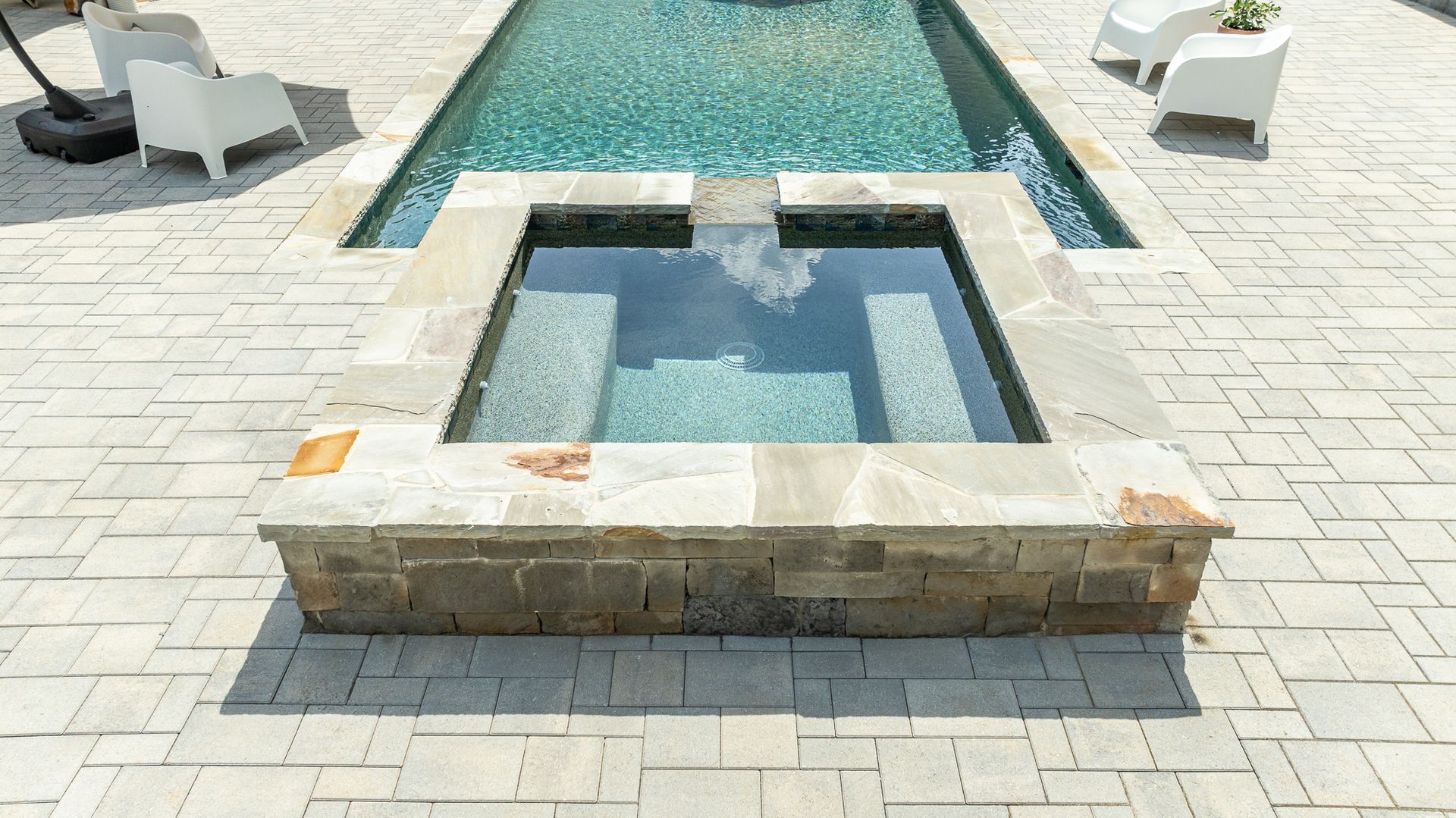 Stone-tiled pool with a small square hot tub in front of a larger swimming pool and white lounge chairs.