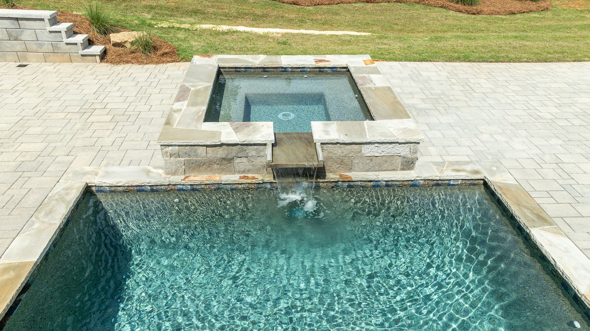 Outdoor pool with a small raised spa and water feature, surrounded by light stone paving and grass.