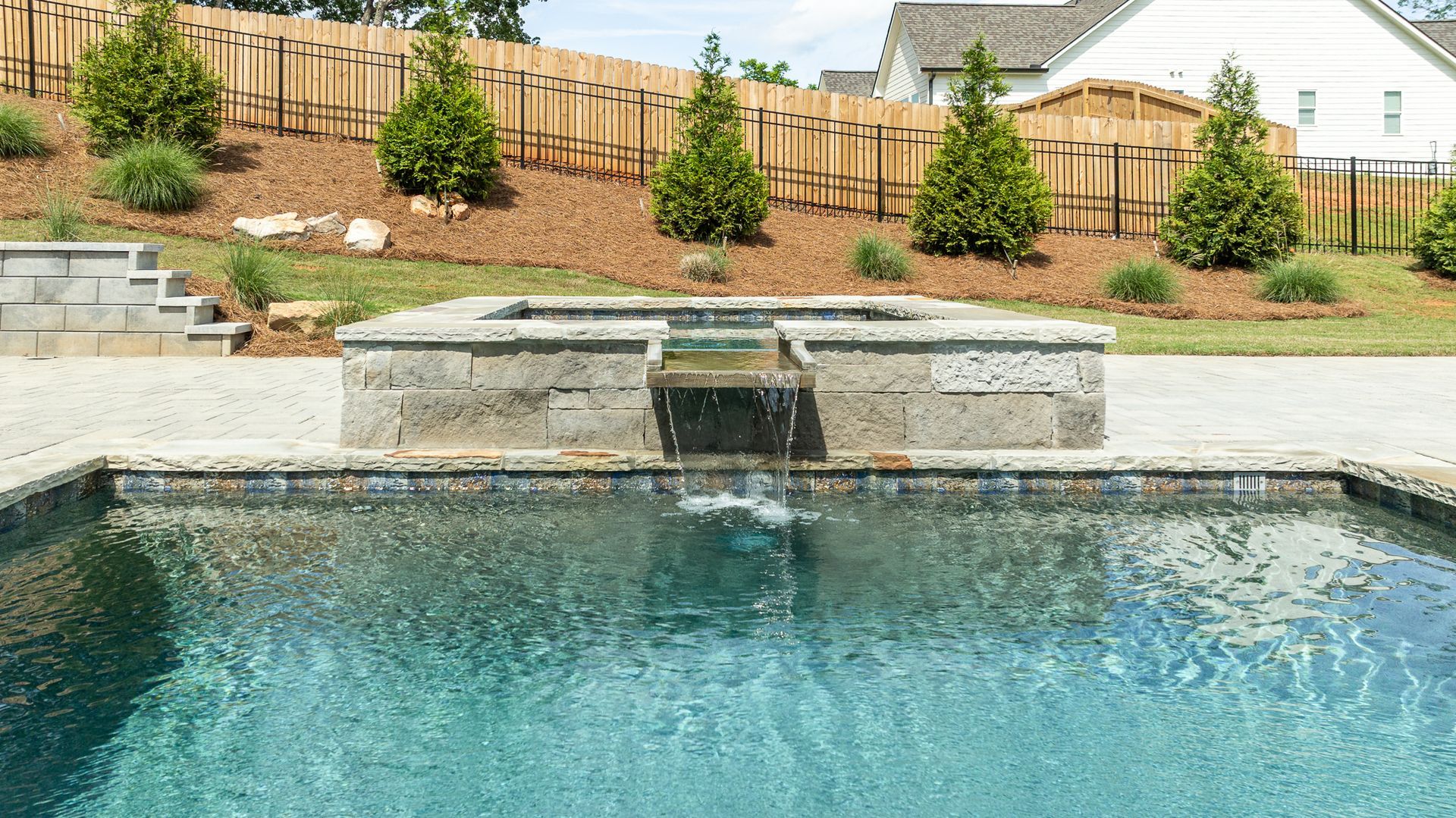 Backyard pool with a stone waterfall, blue water, landscaped retaining wall, and wooden fence.