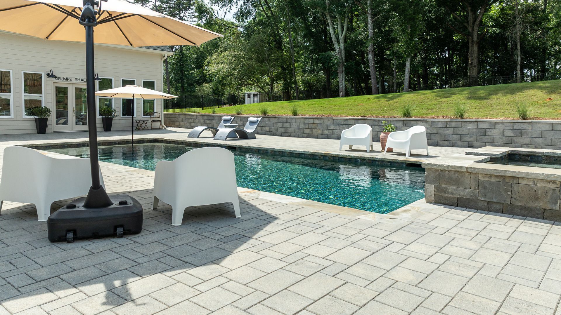Backyard pool with white chairs, umbrella, and stone patio beside a house and trees