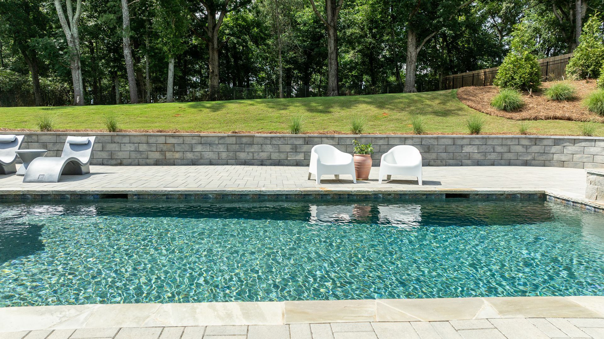 Backyard pool with lounge chairs and a stone wall, framed by lush green trees and shrubs.