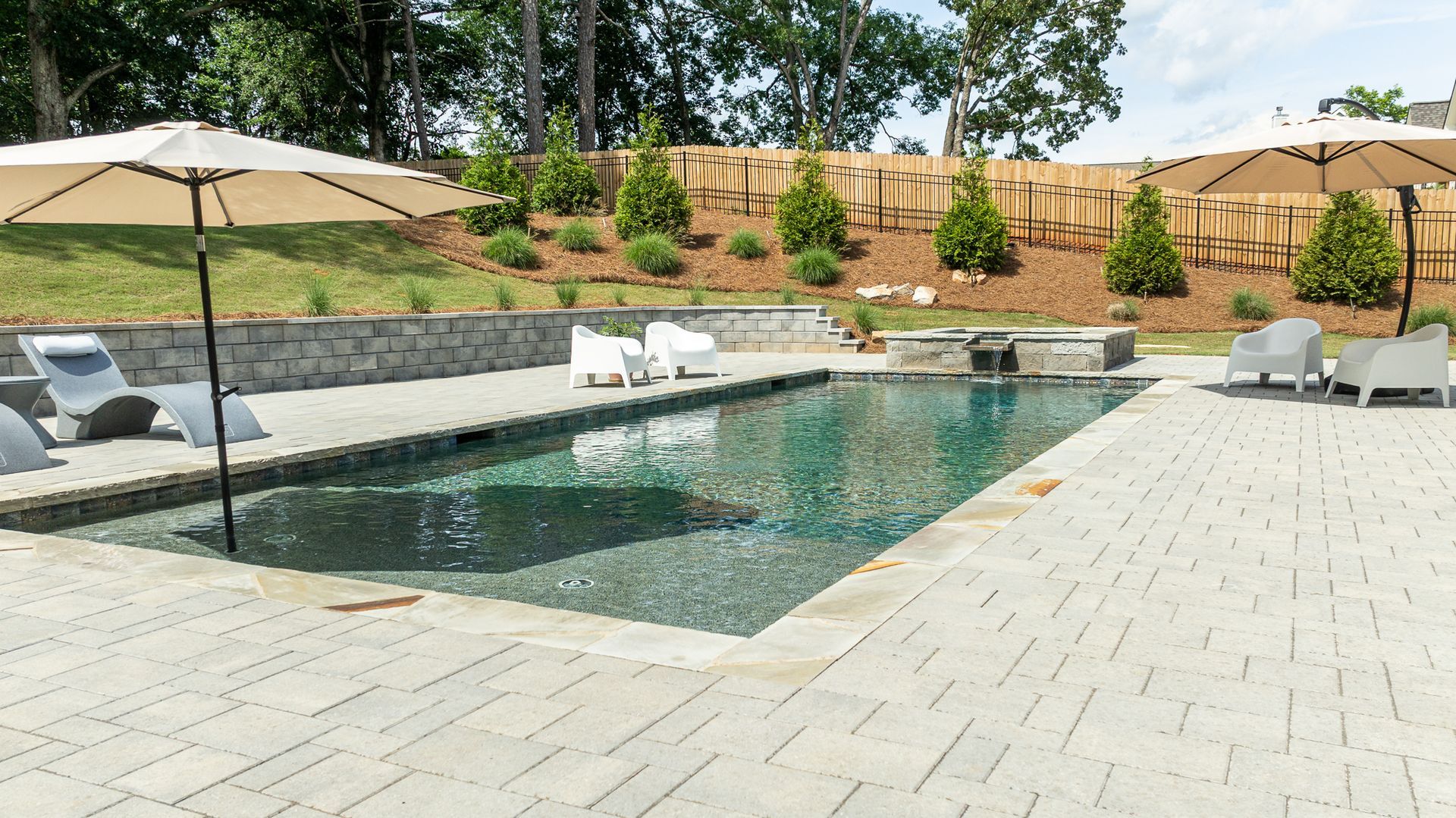 Modern backyard pool with stone patio, lounge chairs, umbrella, and landscaped retaining wall