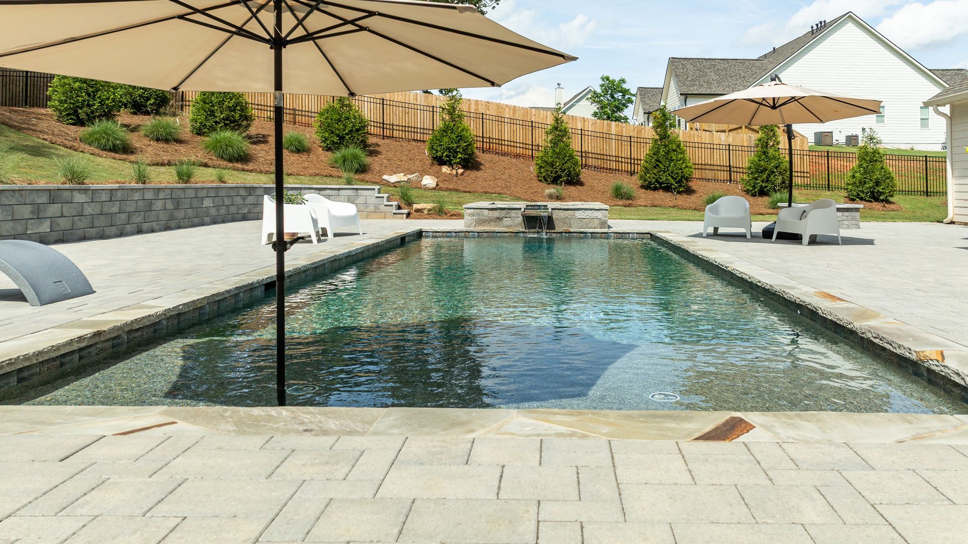 Backyard pool with stone patio, lounge chairs, umbrella, and landscaped fence line