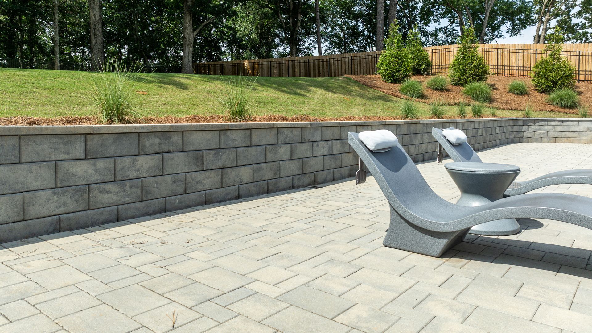 Outdoor patio with two modern gray lounge chairs on a paved stone terrace beside a retaining wall and greenery.