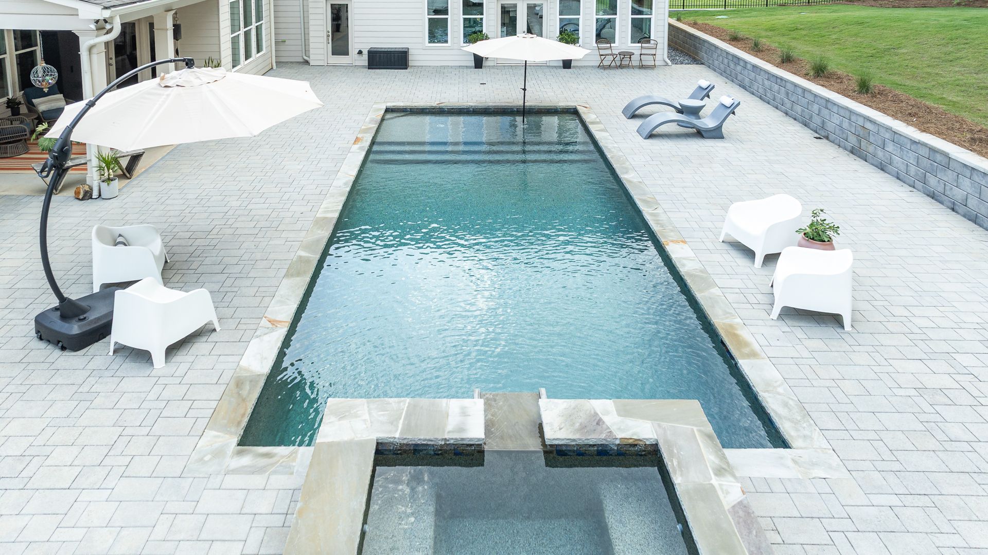 Backyard pool with a small hot tub, white lounge chairs, and umbrellas on a stone patio
