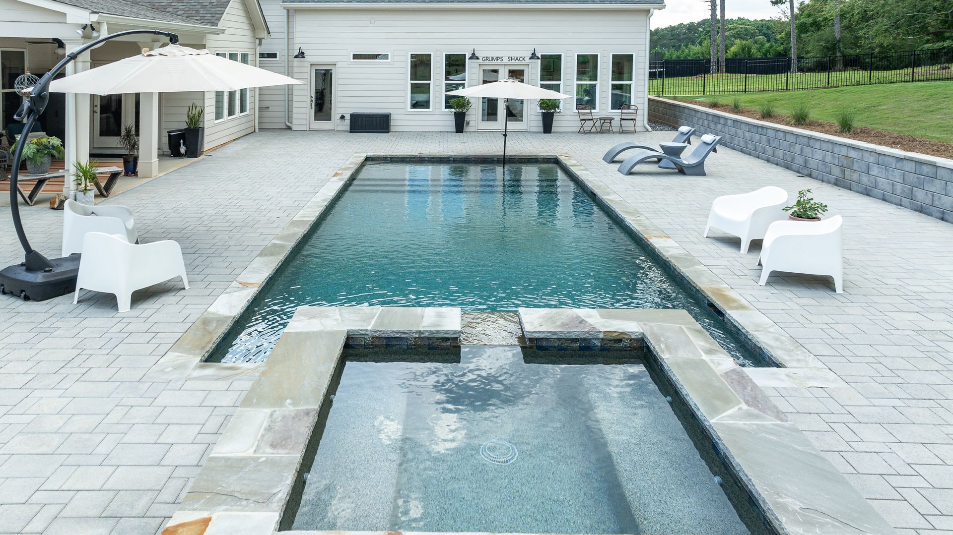 Modern patio with a rectangular swimming pool, hot tub, and white lounge chairs beside a house